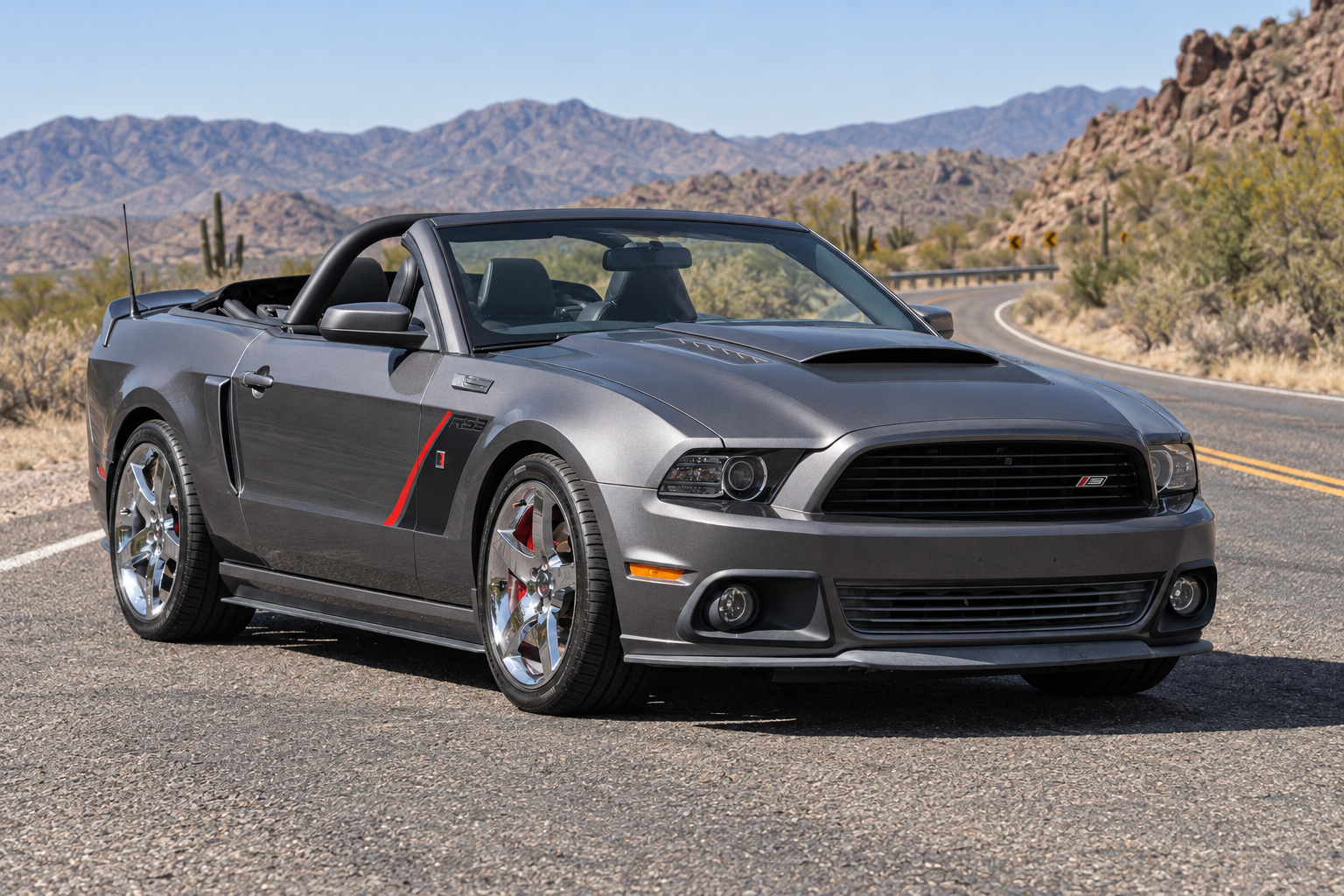 Gray Mustang convertible with a black grille, hood scoop, dark side graphic with a red accent and chrome wheels, parked on a desert road.