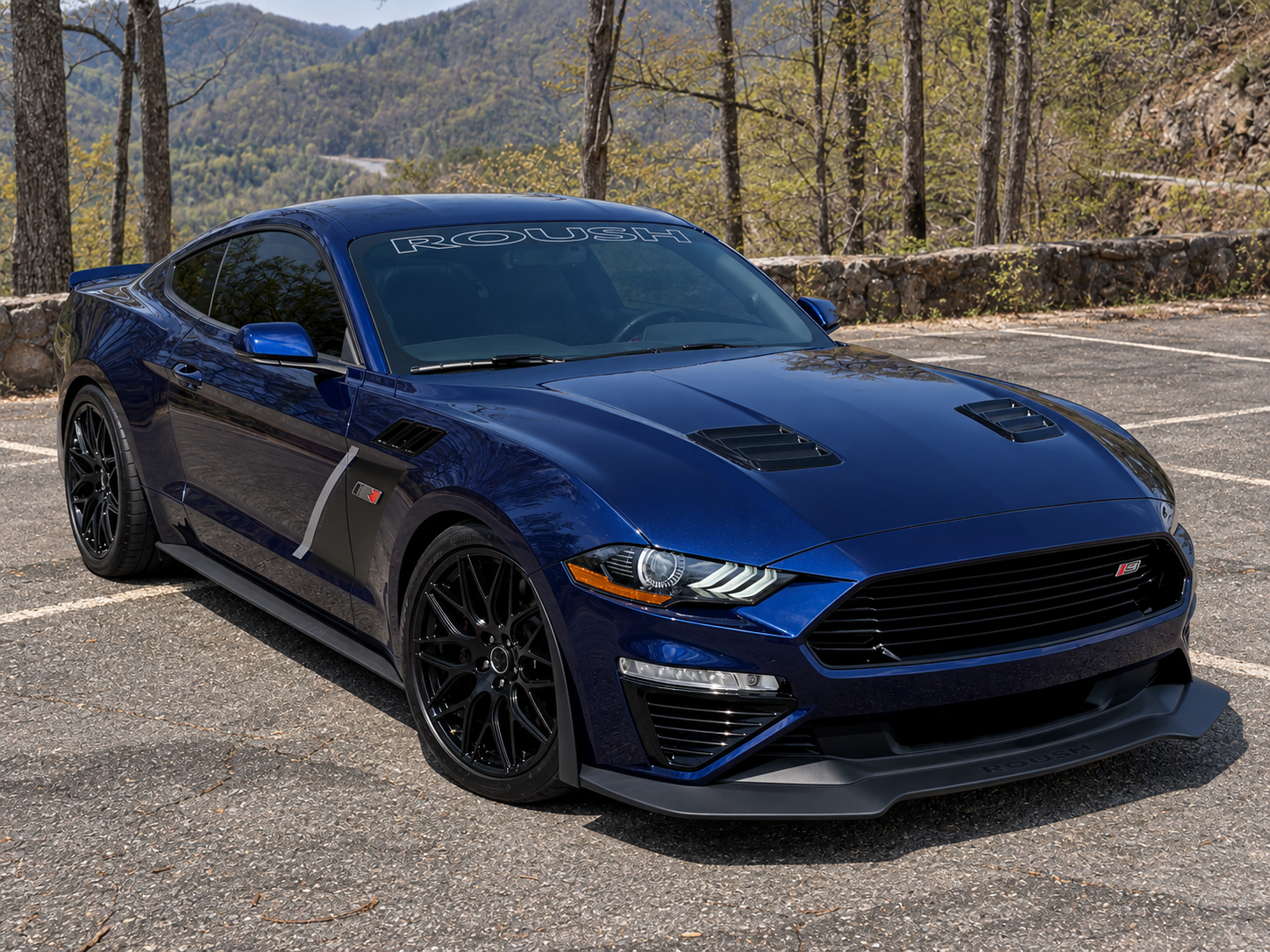 Dark blue Mustang coupe with ROUSH lettering across the windshield, black wheels, hood vents and a front splitter, parked at a wooded overlook.