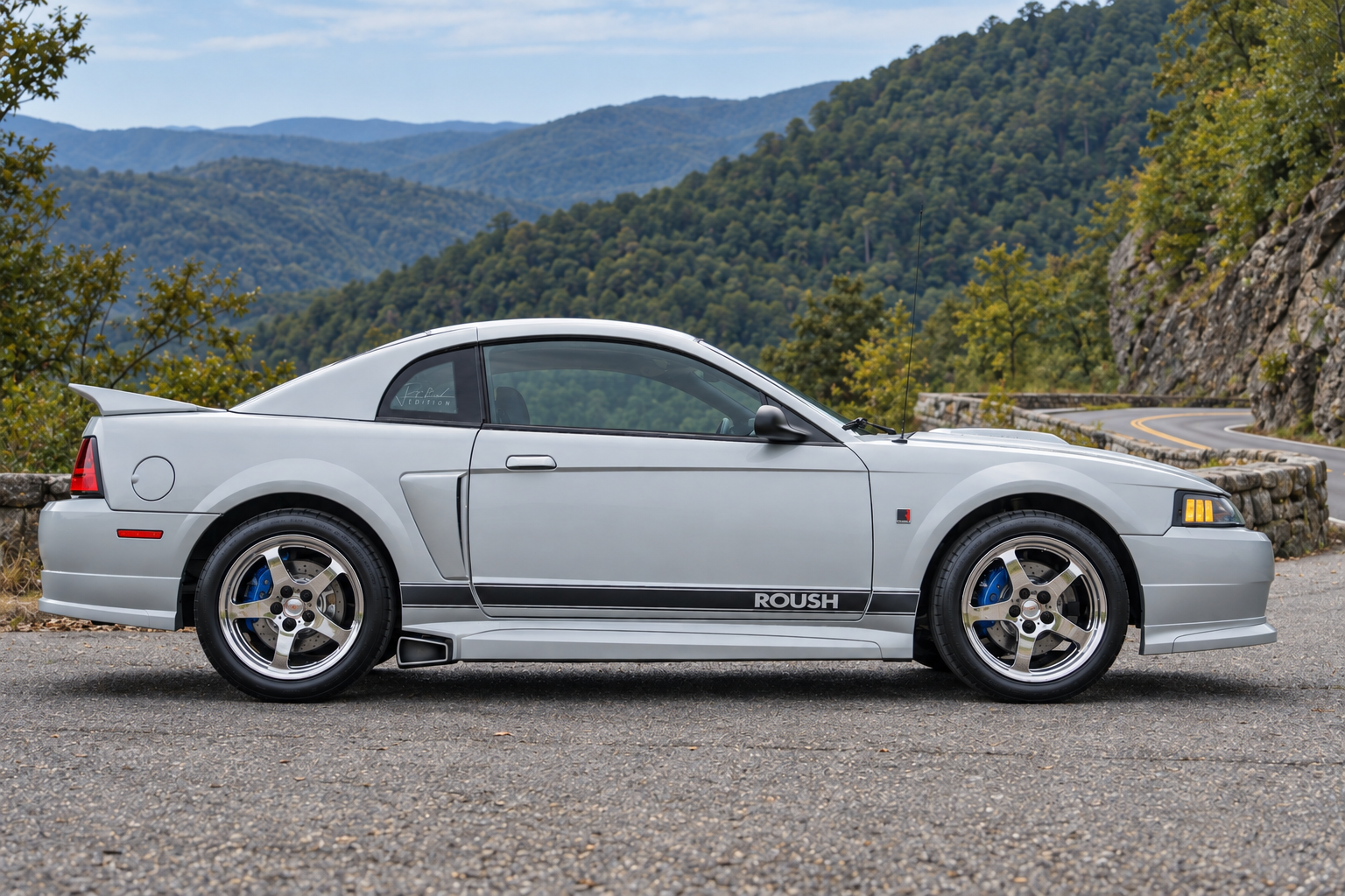 Silver Mustang coupe in profile with ROUSH side-stripe lettering, chrome five-spoke wheels and blue brake calipers, on a mountain overlook.